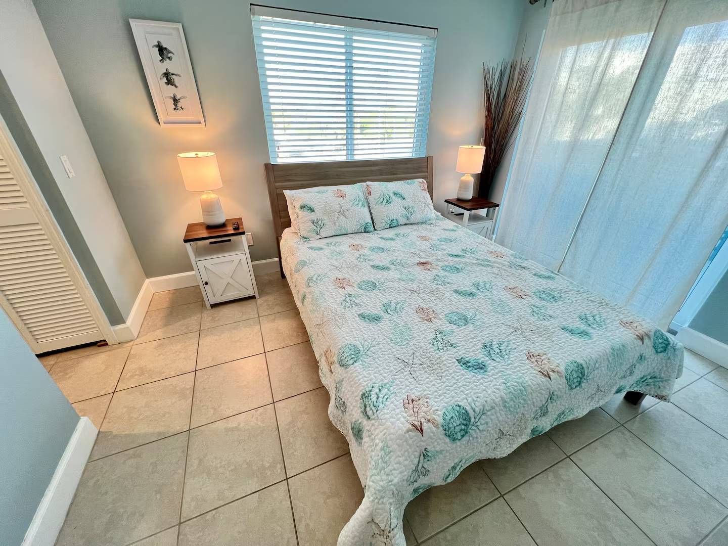A bedroom with a bed, bedside tables, a window with blinds, and a door.