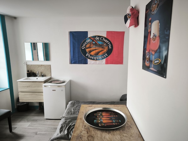Une petite chambre moderne, dotée d’un lit simple, d’une table de chevet avec un plateau contenant des articles de toilette, d’un petit lavabo blanc et d’un réfrigérateur compact.