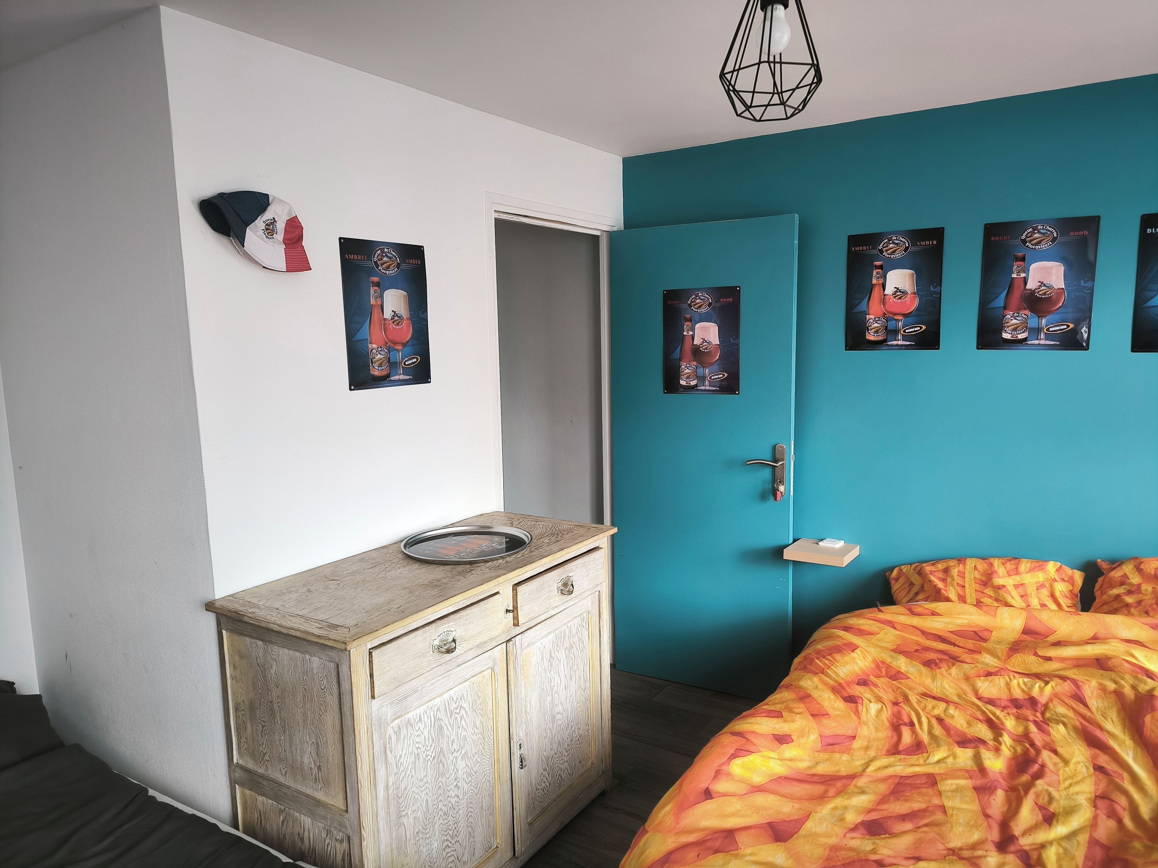 A bedroom with a teal accent wall, a wooden cabinet with a glass dish, and a bed with a patterned blanket.