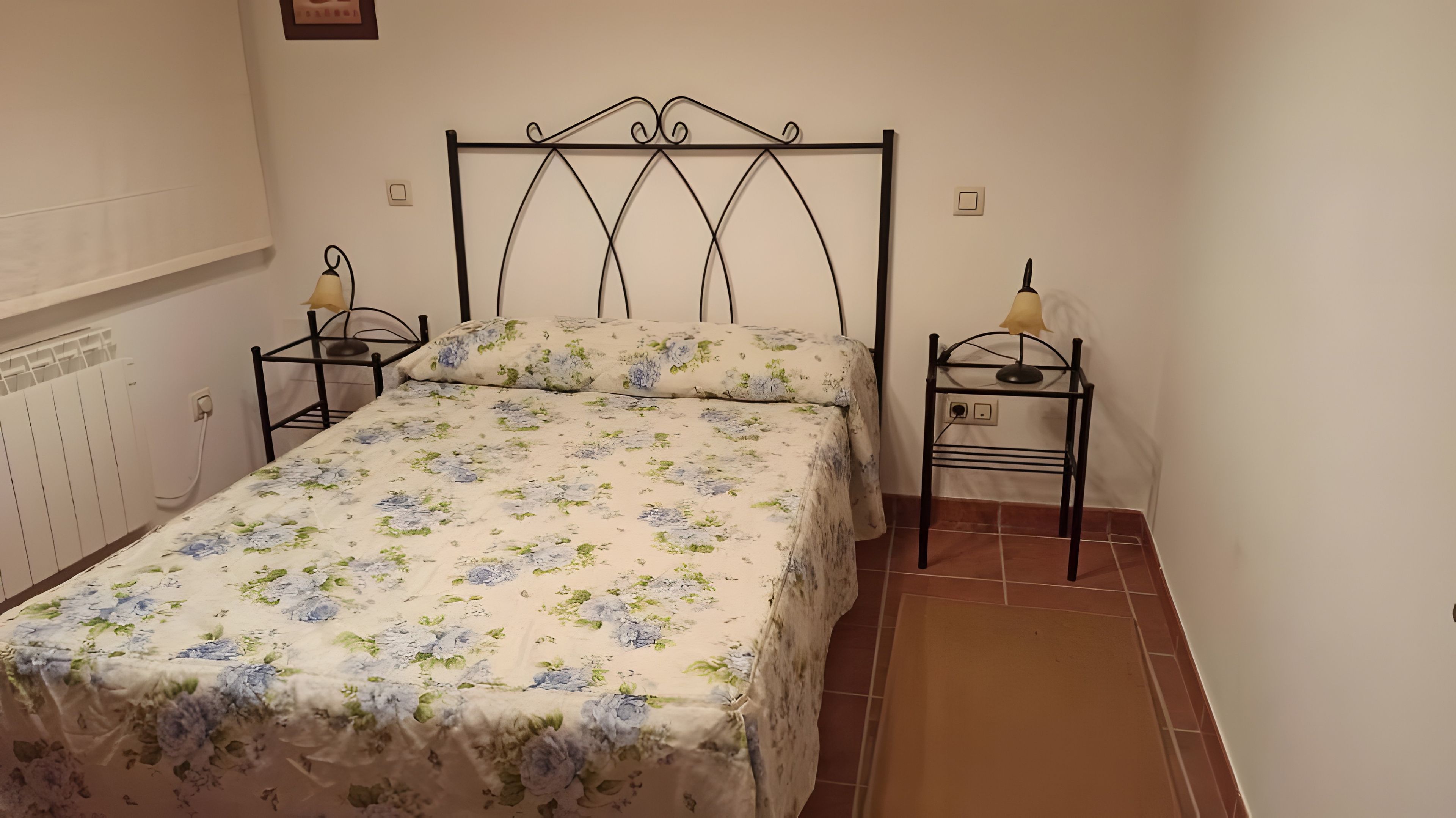 Una cama individual con una colcha estampada, dos mesitas de noche con lámparas, un radiador y suelo de baldosas.