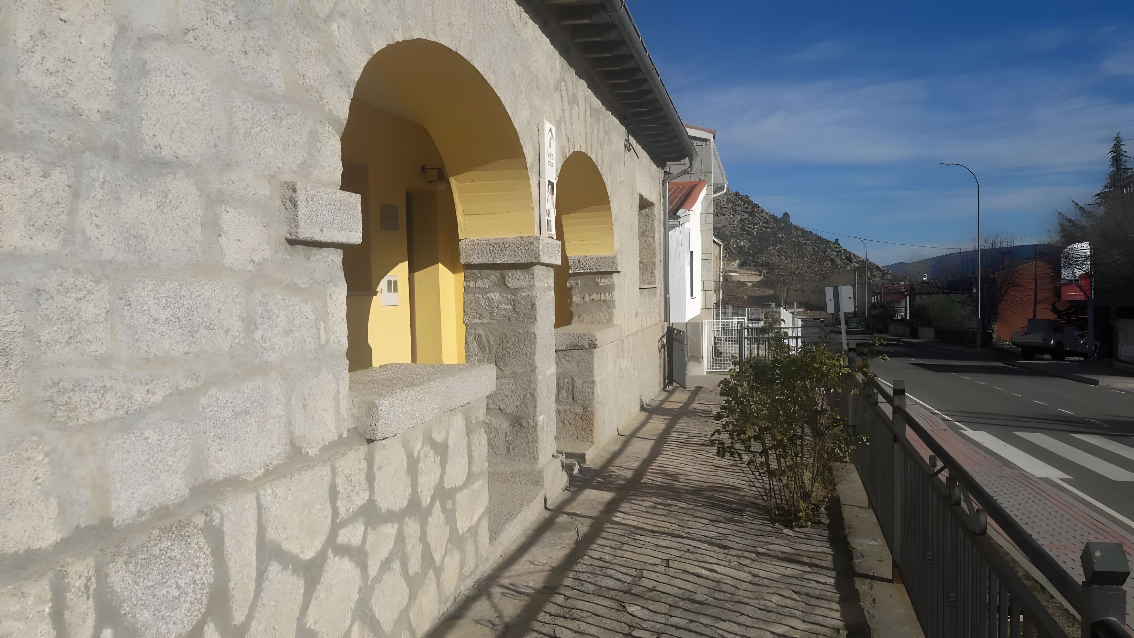 Un edificio de muros de piedra y arcos amarillos, un sendero pavimentado y una calle con vehículos.