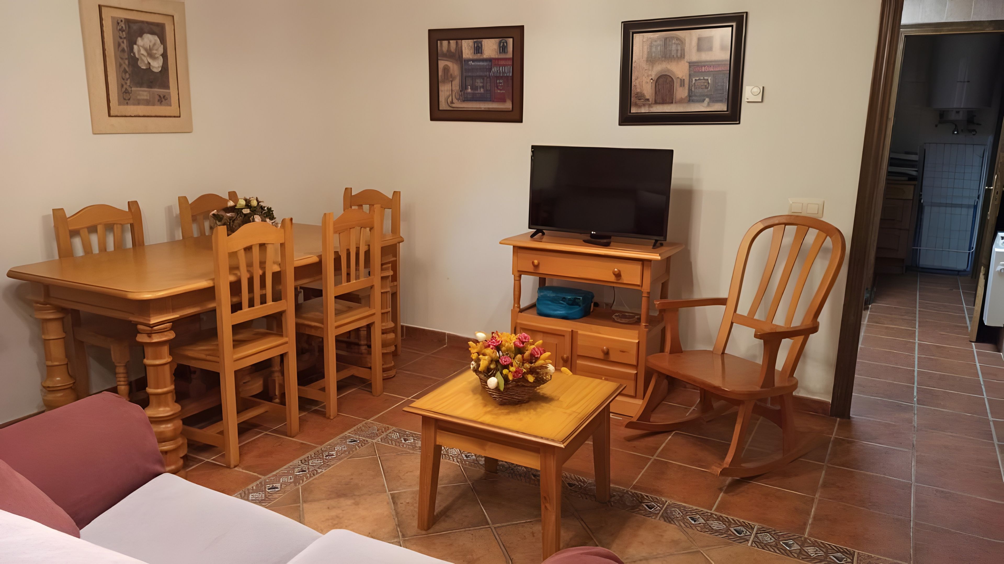 Una sala de estar con una mesa de comedor de madera, sillas, un televisor sobre un mueble de madera, un sofá y una cesta decorativa en el suelo.