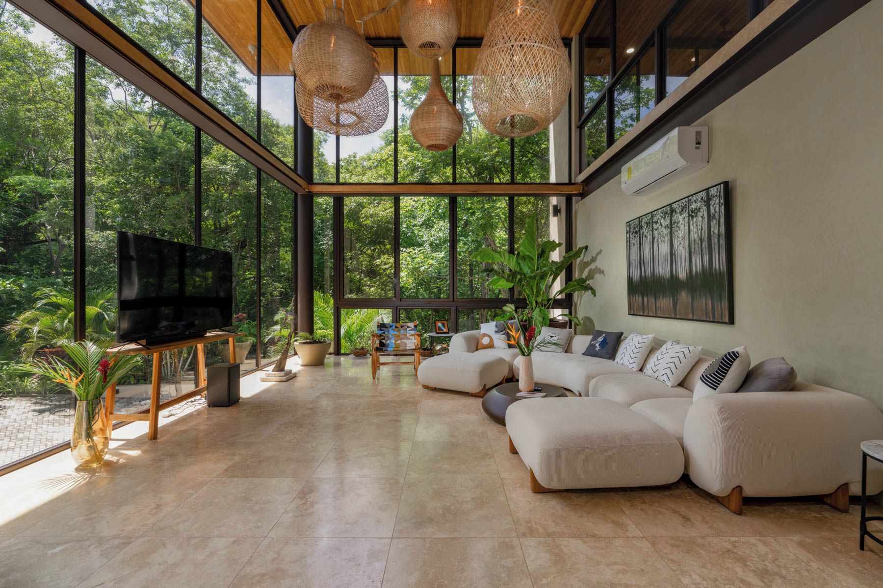 A spacious living room with a large sectional sofa, a television, and a view of lush greenery through large windows.