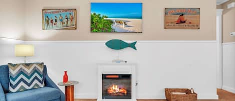 Uma sala de estar com lareira, um sofá azul, uma mesinha de centro com abajur e quadros com uma cena de praia.