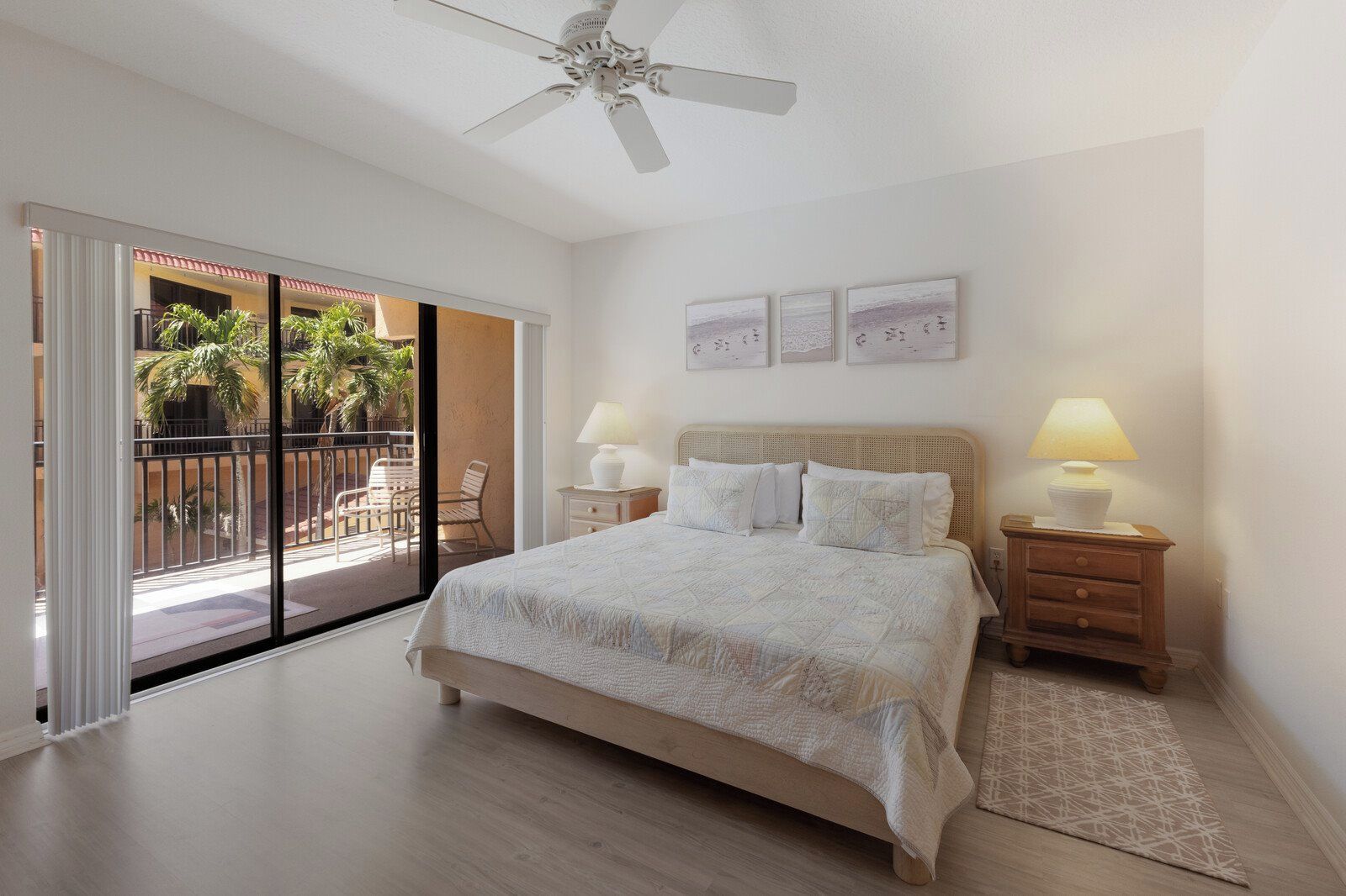 Une chambre à coucher comprenant un lit, deux tables de chevet avec des lampes, un ventilateur de plafond et une vue sur un balcon aménagé avec du mobilier d’extérieur.