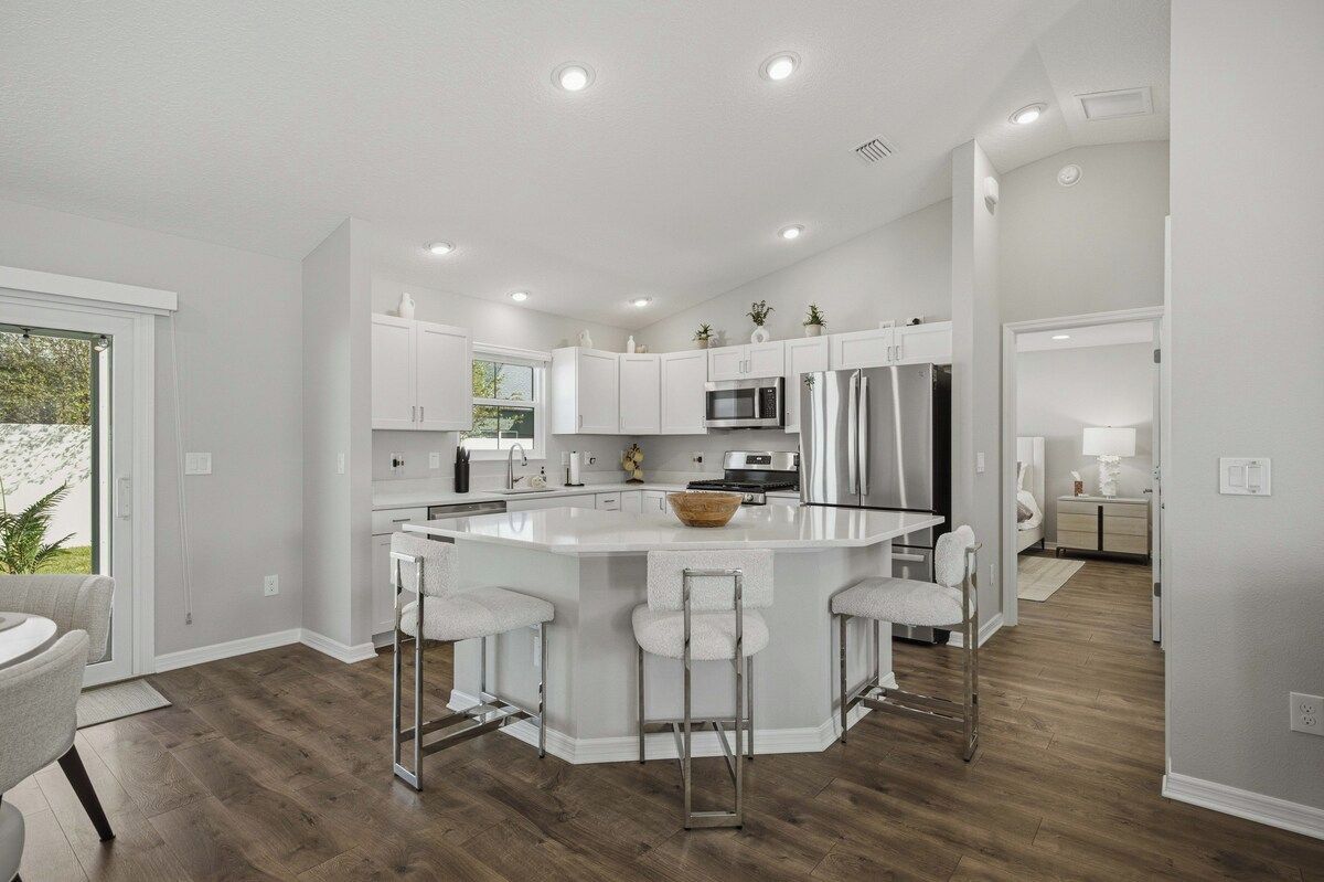 A modern kitchen with white cabinetry, a central island with bar stools, stainless steel appliances, and recessed ceiling lighting.