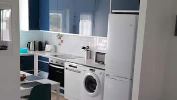 Een kleine keuken met een wasmachine, een eethoek met een tafel en stoelen, en een zithoek met een bank.