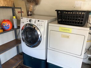 Ein Waschbereich mit einer Frontlader-Waschmaschine und einem Trockner, einem Wäschekorb auf dem Trockner und Reinigungsmitteln in einem Regal.