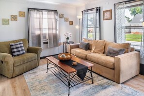 Ein Wohnzimmer mit Sofa, Ohrensessel, Couchtisch und Blick ins Freie.