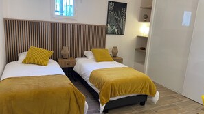 A hotel room with two beds, each with a yellow throw blanket and a patterned pillow. There is a window with a view of a building, a wall-mounted plant, and a shelf with a globe and other items. - New Listing (Les Pennes-Mirabeau)