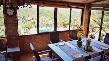 Un coin repas avec une table dressée pour deux personnes, de grandes fenêtres donnant sur un paysage vallonné et un mur en briques.