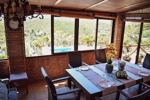 Un coin repas avec une table dressée pour deux personnes, de grandes fenêtres donnant sur un paysage vallonné et un mur en briques.