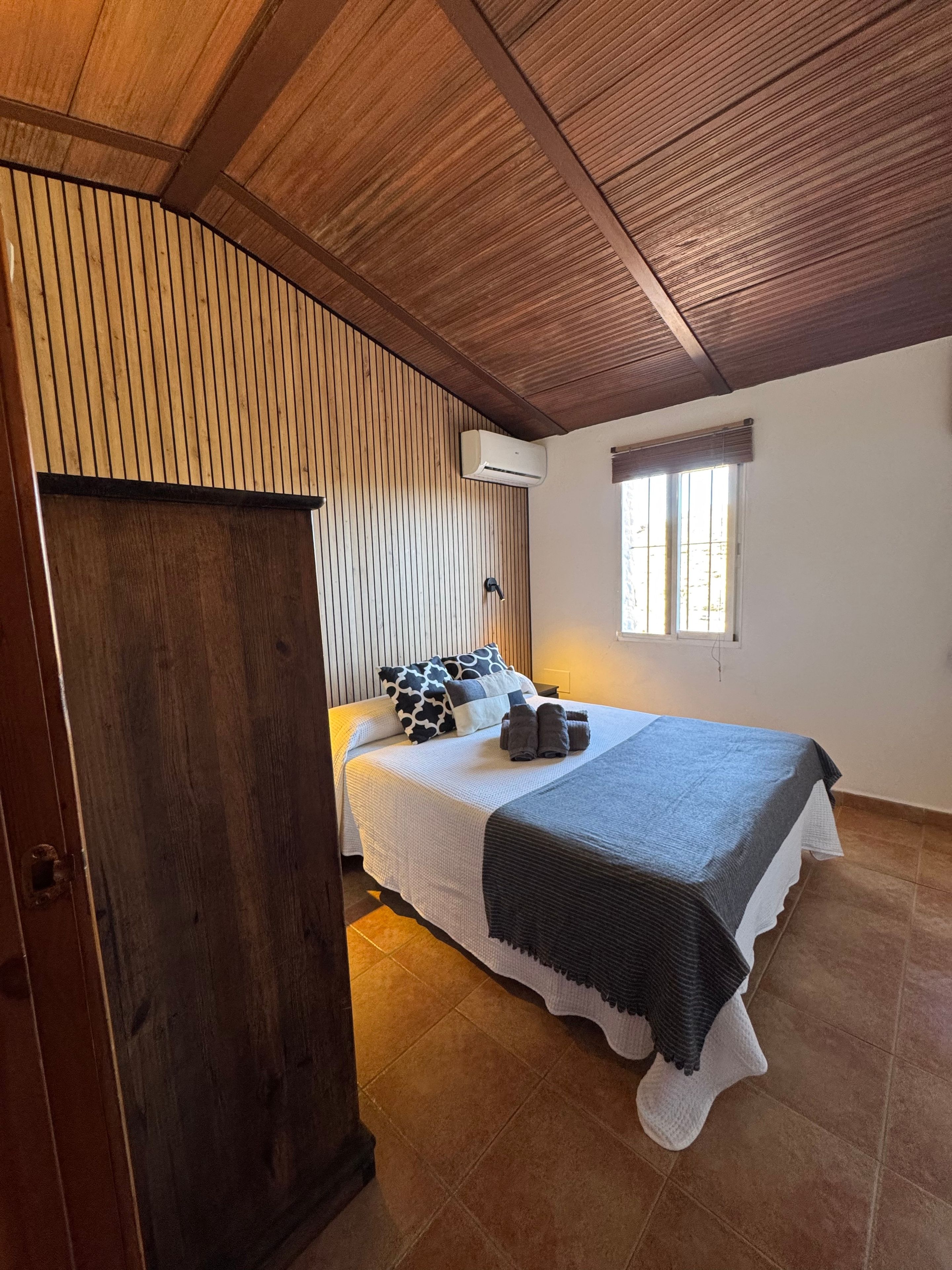 Une chambre avec un plafond en bois, un lit recouvert d’une courtepointe blanche et grise, une fenêtre avec des stores et une porte.