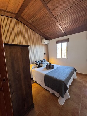 Une chambre avec un plafond en bois, un lit recouvert d’une courtepointe blanche et grise, une fenêtre avec des stores et une porte.