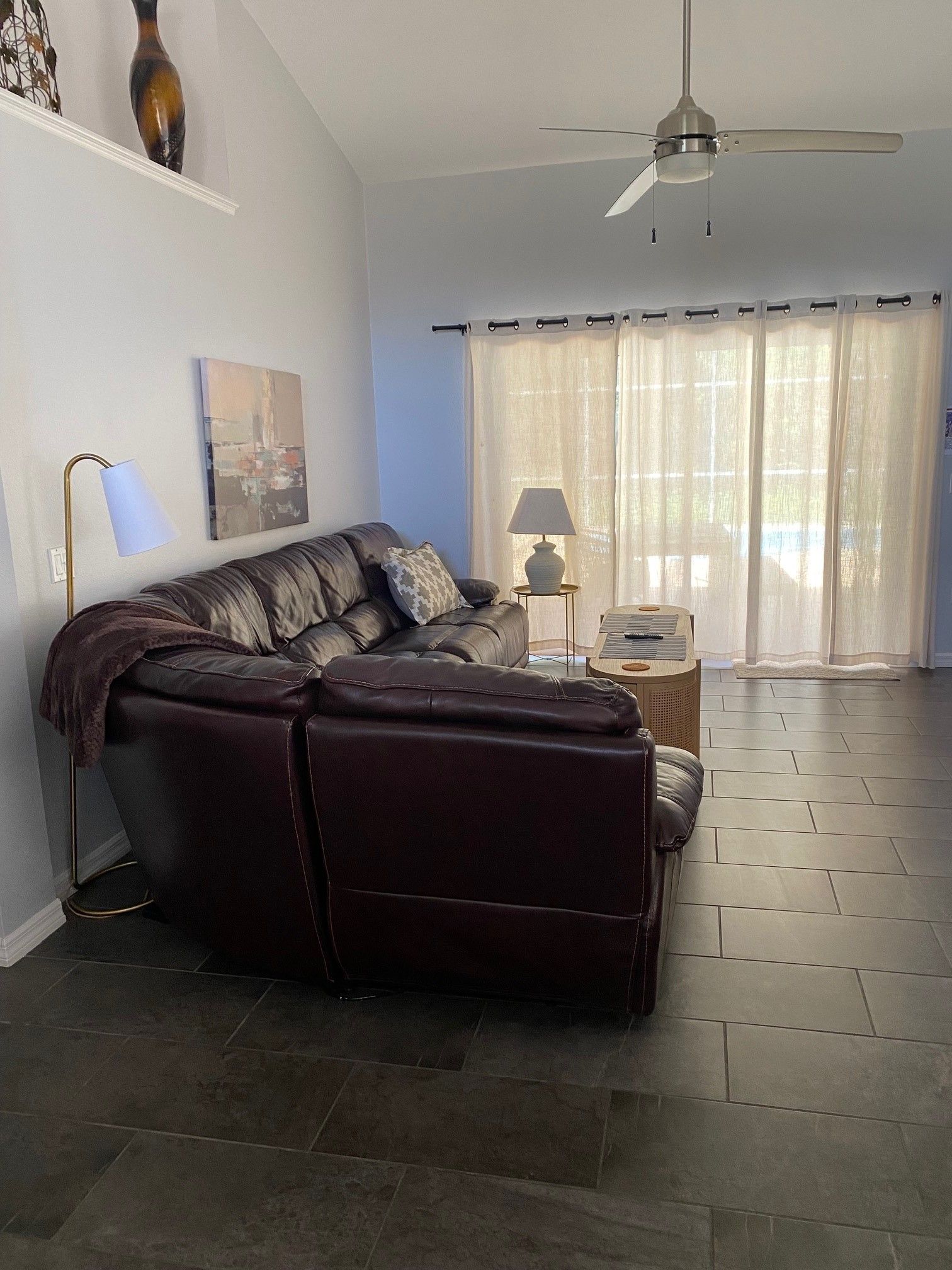 Sala de estar com um sofá de couro marrom escuro, luminária de chão, mesa com controle remoto e ventilador de teto.