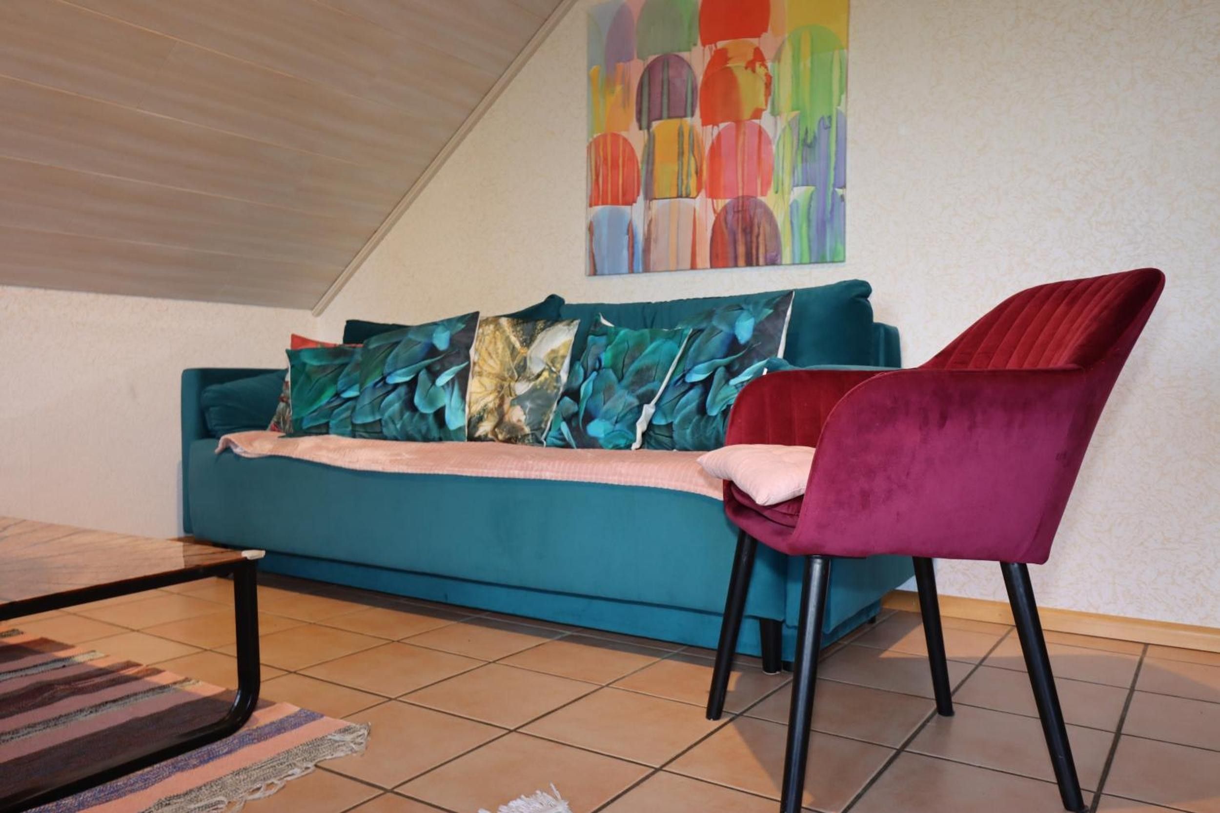 A teal sofa with patterned cushions, a red velvet chair, and a colorful abstract painting on the wall.
