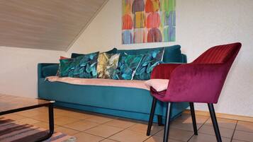 A teal sofa with patterned cushions, a red velvet chair, and a colorful abstract painting on the wall.
