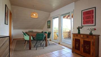A dining area with a wooden table and chairs, a hanging light fixture, a sliding glass door leading to an outdoor deck, and a cabinet with floral designs.