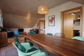 En modern inredning med ett matbord i trä, gröna stolar, en flätad lampa och ett köksområde.