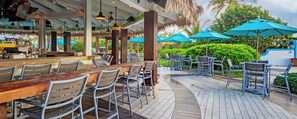 Un área de bar al aire libre con mesas de madera y sillas de metal, rodeada de sombrillas y follaje.