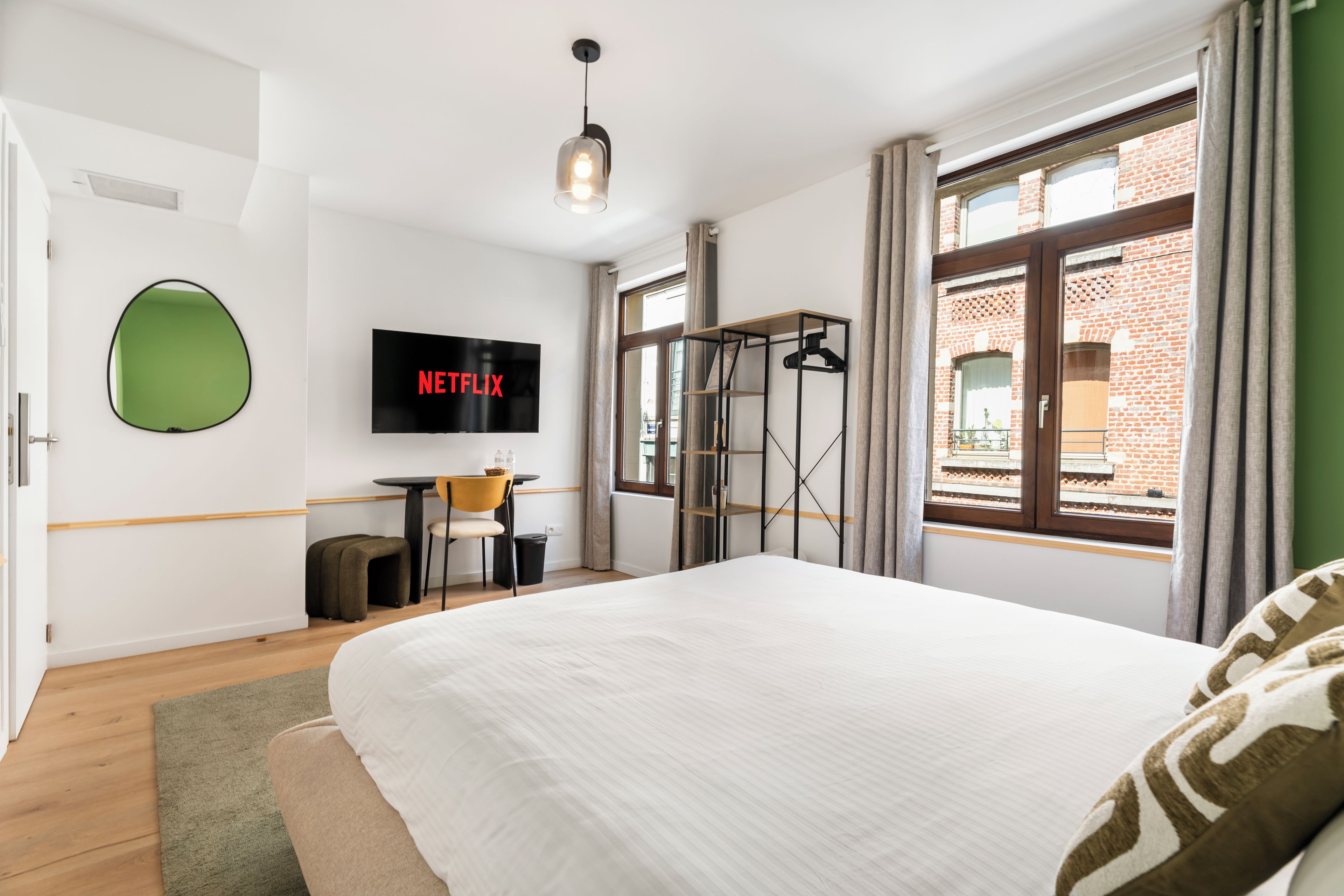 A modern hotel room with a large bed, a flat-screen TV mounted on the wall, a small desk with a chair, and a window with a view of a brick building.