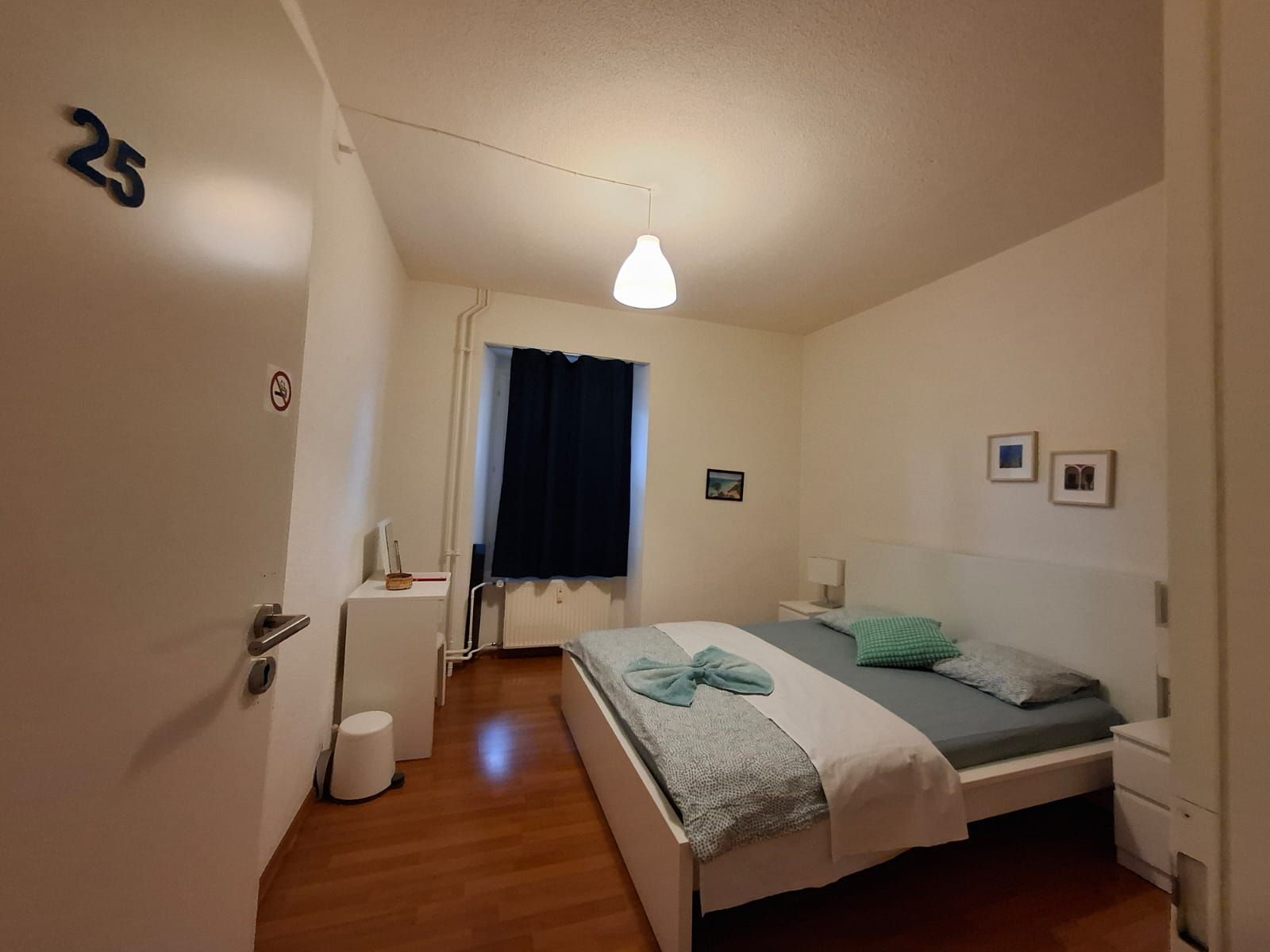 Un dormitori amb un llit, taules de nit, una finestra amb cortines, una làmpada penjada a la paret i una porta amb un número.