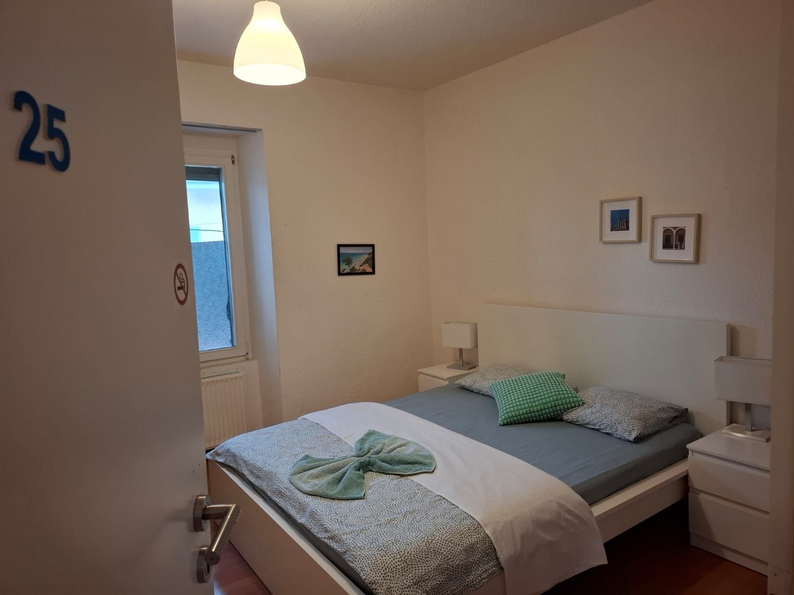 Un dormitori amb un llit, una tauleta de nit, una làmpada penjada a la paret, una porta amb un número i una finestra.