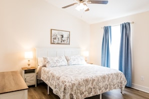 Une chambre à coucher comprenant un lit, deux tables de chevet, un ventilateur de plafond, une fenêtre avec des rideaux et un tableau accroché au mur.