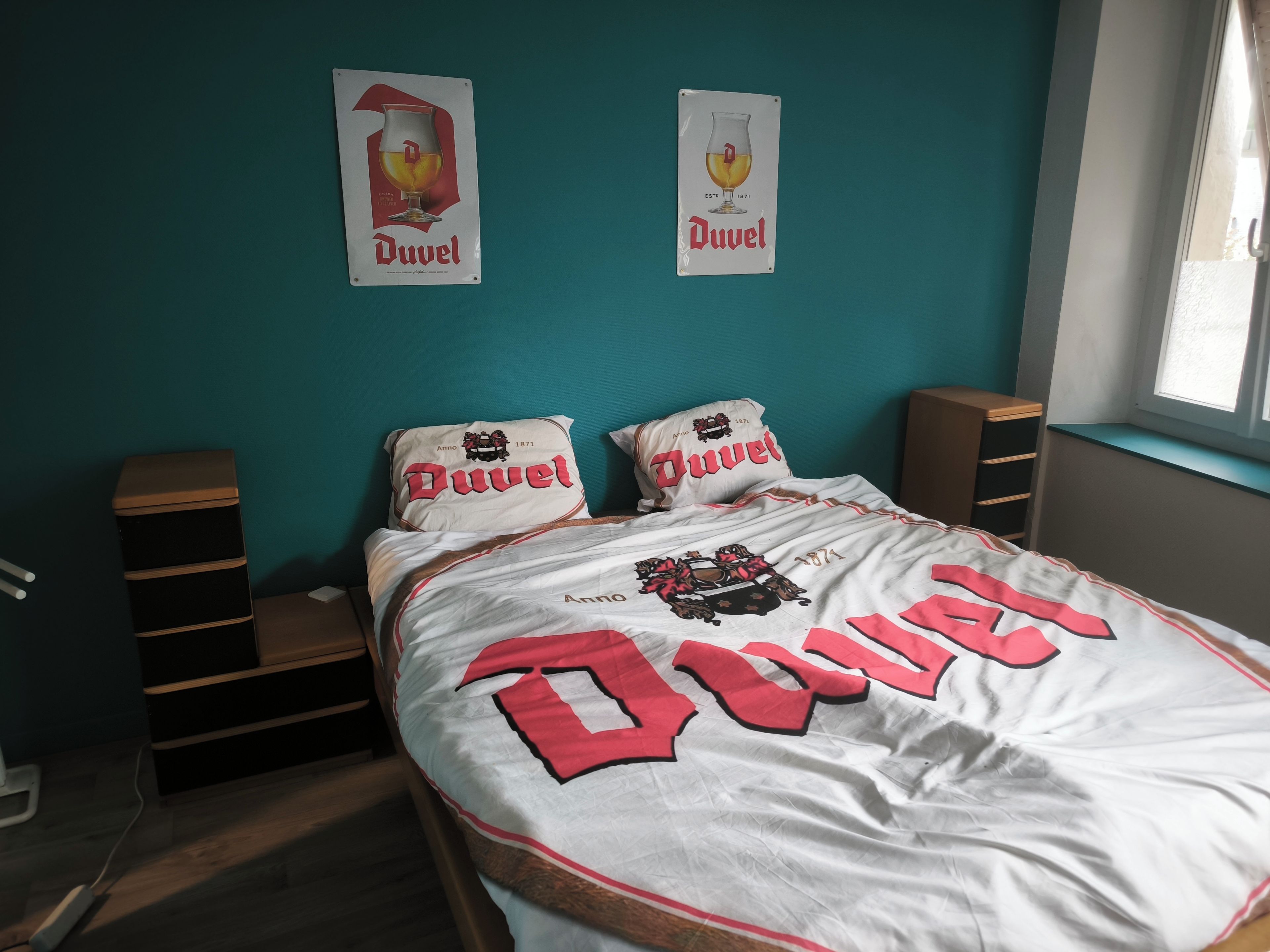 A bed with a duvet featuring the Duvel logo, two pillows with the same logo, and two wall-mounted posters of a beer glass with the Duvel logo.