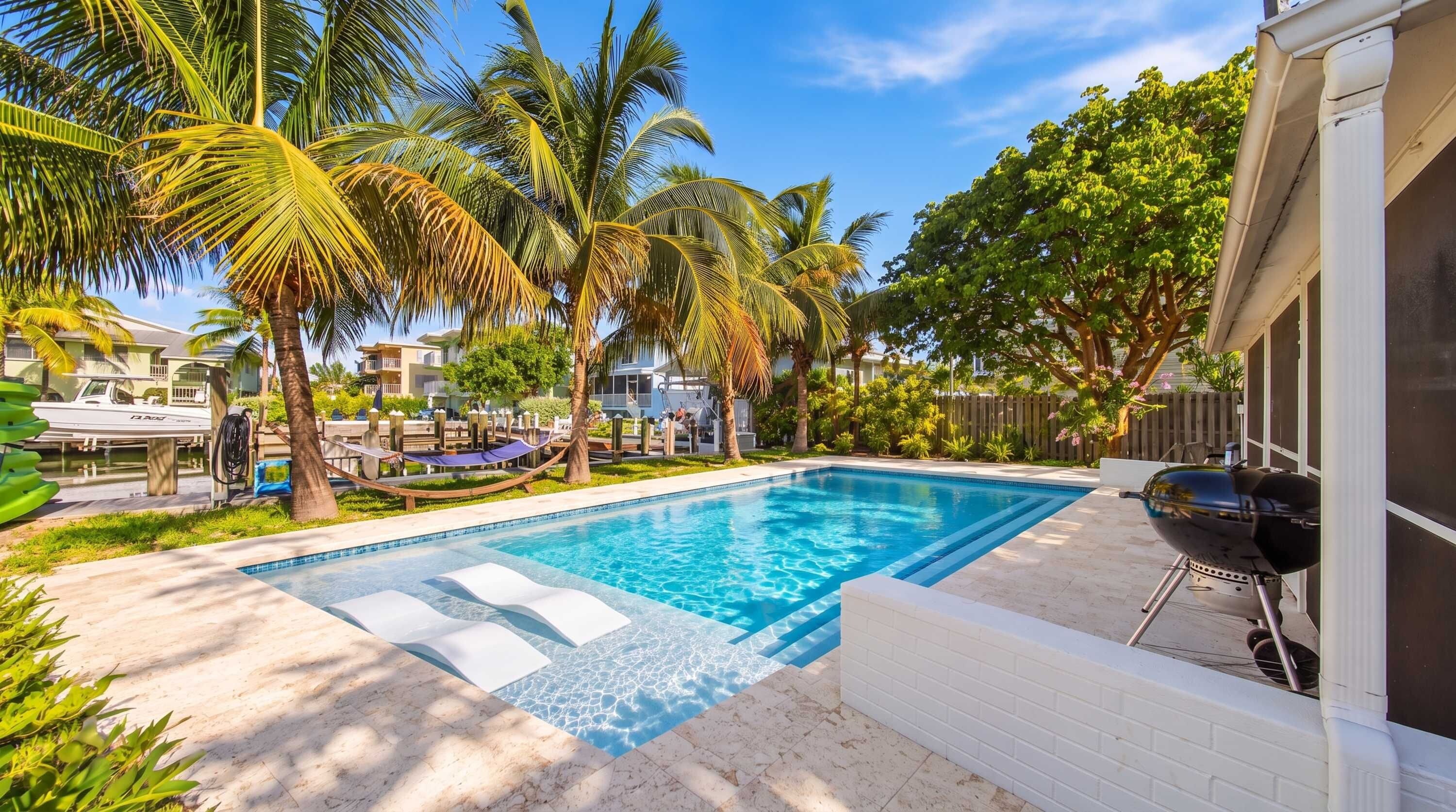 A swimming pool with a surrounding deck, palm trees, and a barbecue grill.
