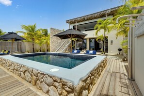 En swimmingpool med stenvægge, en træterrasse og et moderne hus med stenfacade og en trappe op til en balkon.