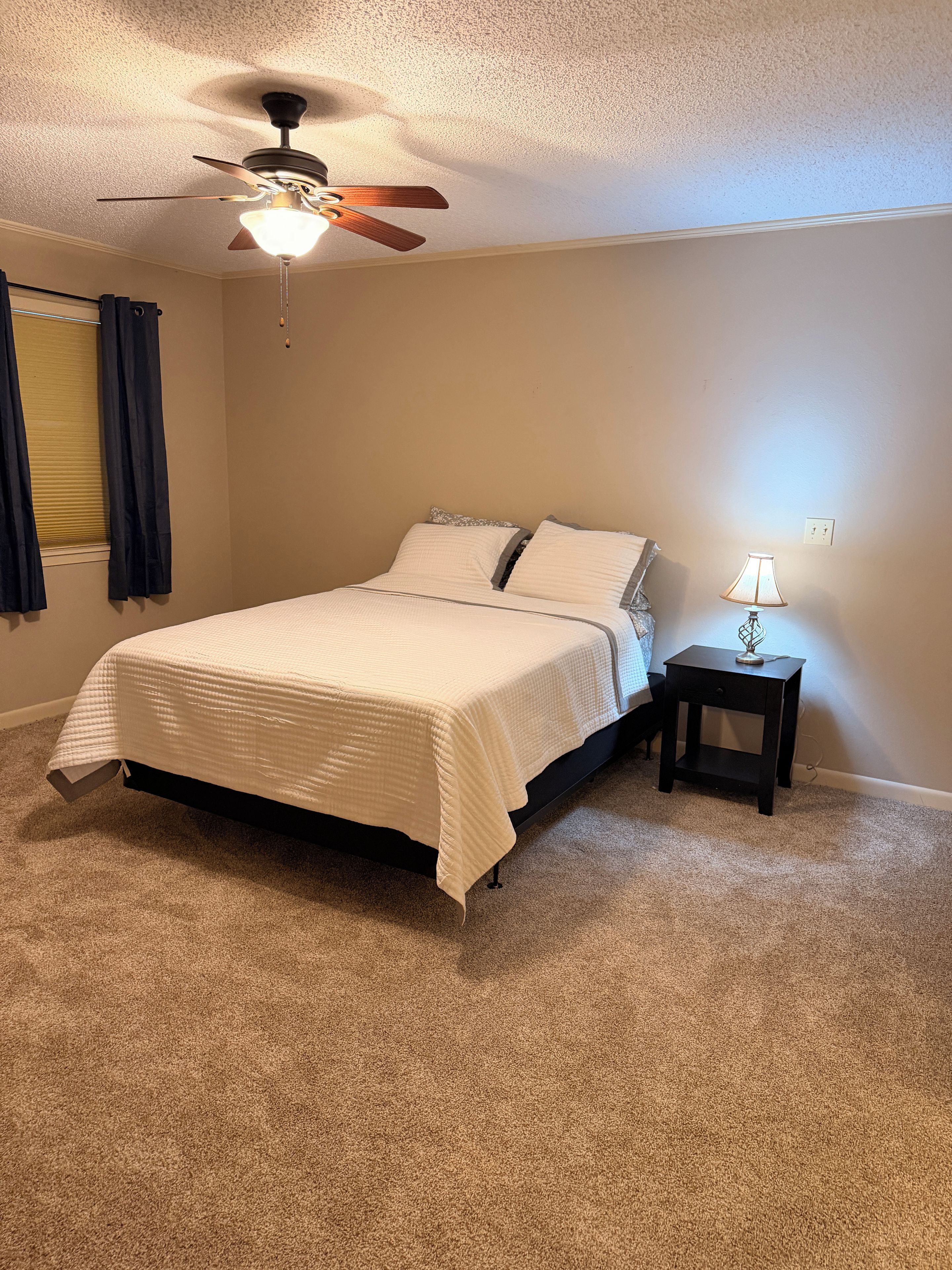 A bedroom with a bed, a nightstand with a lamp, a ceiling fan, and a window with curtains.