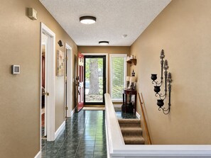 A hallway with a door leading to a bright outdoor area, a staircase with a handrail, and a wall-mounted light fixture. - Esmeralda Escape In the Woods, Close to the Fun (Hot Springs)