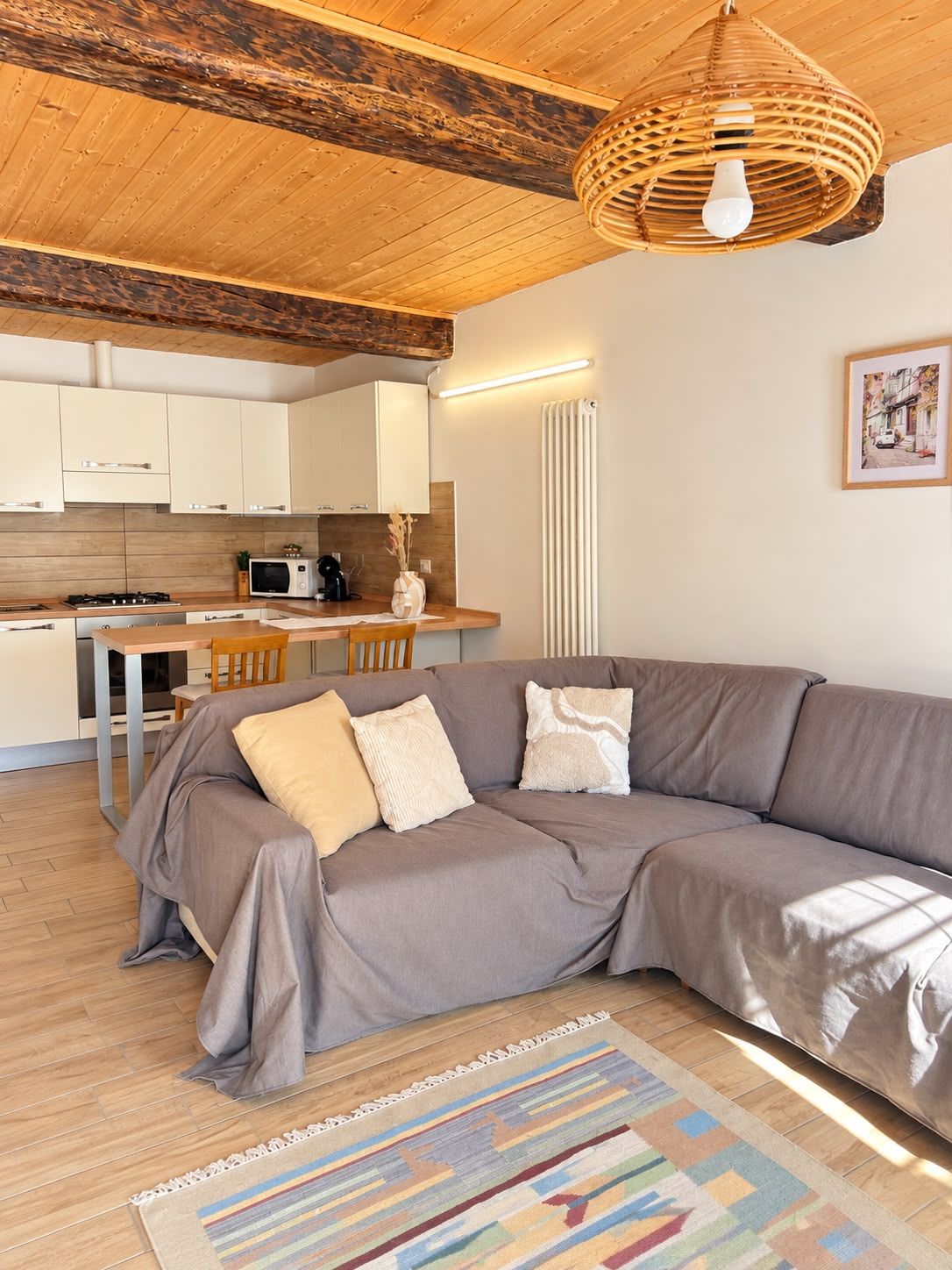 Un espai de viure modern amb un sofà gris, terres de fusta, una làmpada de sostre teixida i una zona de cuina amb armaris blancs i un forn elèctric.
