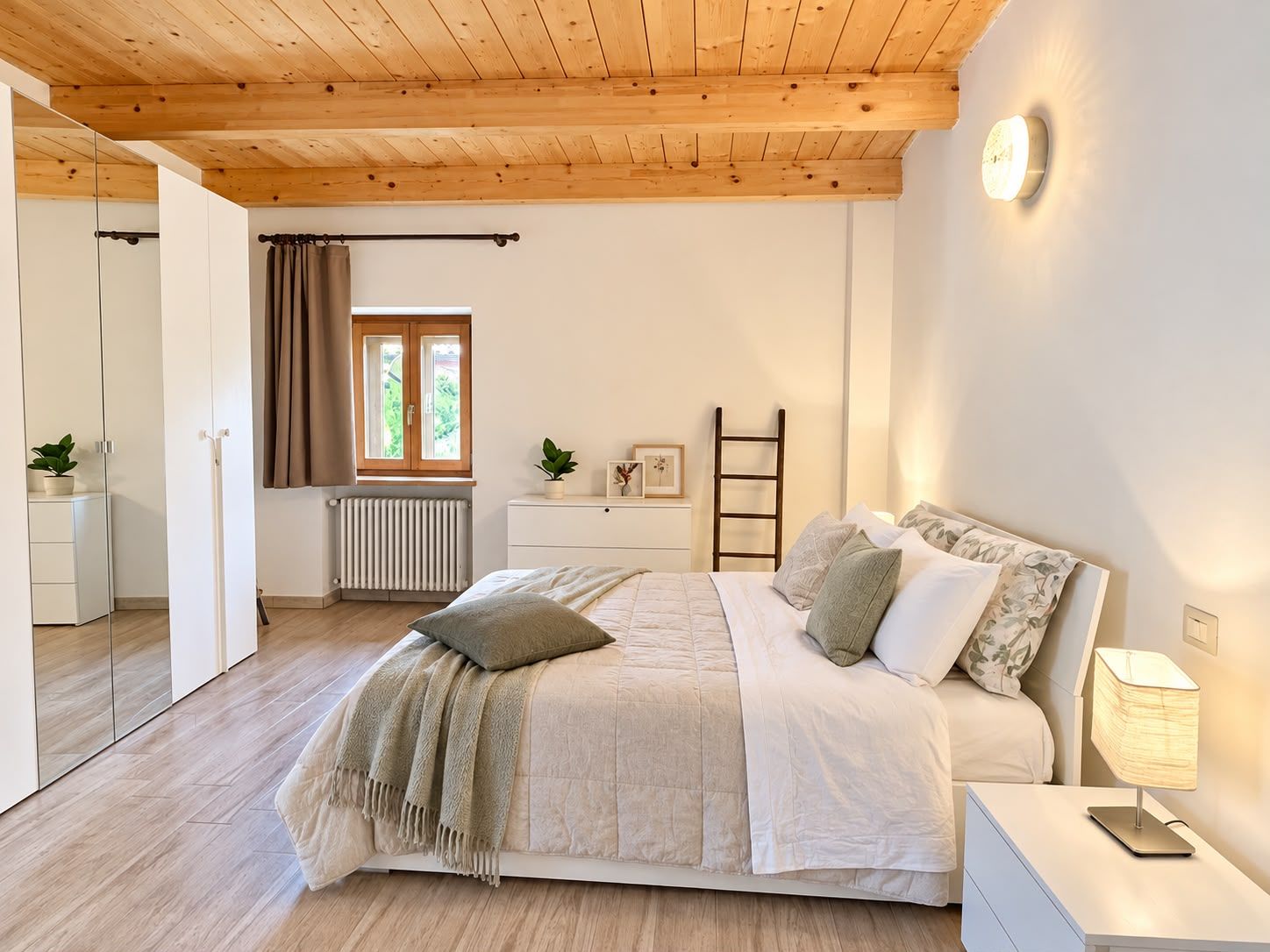 Un dormitorio con techo de madera, una cama con manta color beige, una mesita de noche con lámpara y una ventana con cortinas.