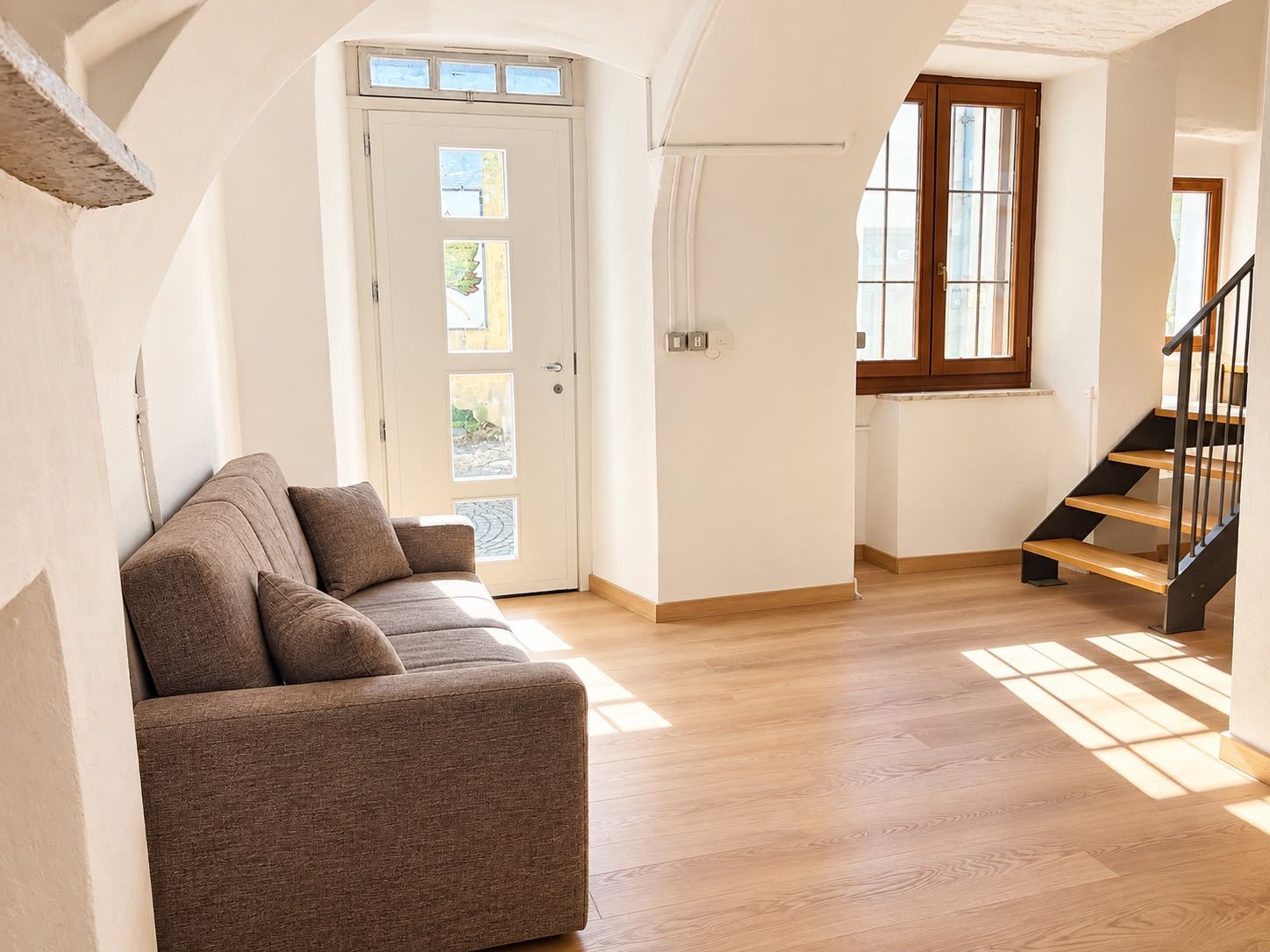 Una habitación amplia con piso de madera, un sofá, una puerta con paneles de vidrio y una escalera con escalones de madera.