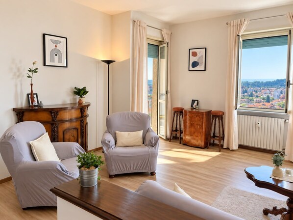 A modern living room with two grey armchairs, a wooden sideboard, a floor lamp, and a view of a cityscape through a large window. - QuickHome Panorama - Two Bedroom Apartment, Sleeps 4 (Cossato)