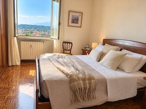 A hotel room with a large bed, a view of a town through a window, a radiator, a wooden chair, and a bedside table with a lamp. - QuickHome Panorama - Two Bedroom Apartment, Sleeps 4 (Cossato)