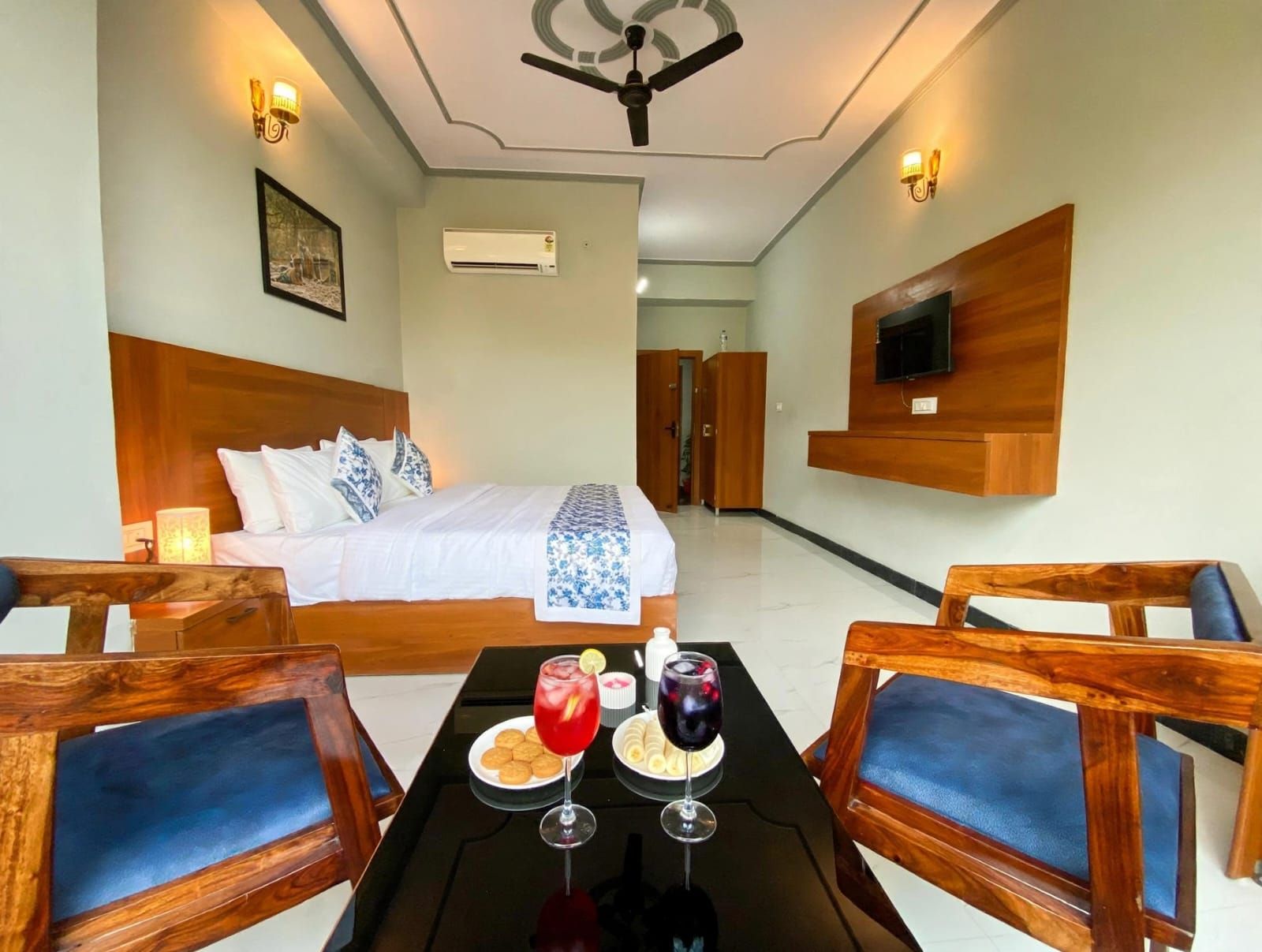 A hotel room with a bed, a TV mounted on the wall, a table with drinks and snacks, and a ceiling fan.