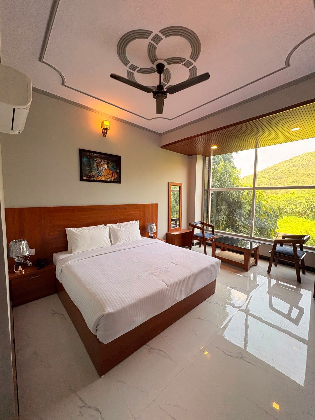 A hotel room with a large bed, a ceiling fan, a painting, a nightstand, a desk, and a view of greenery outside.