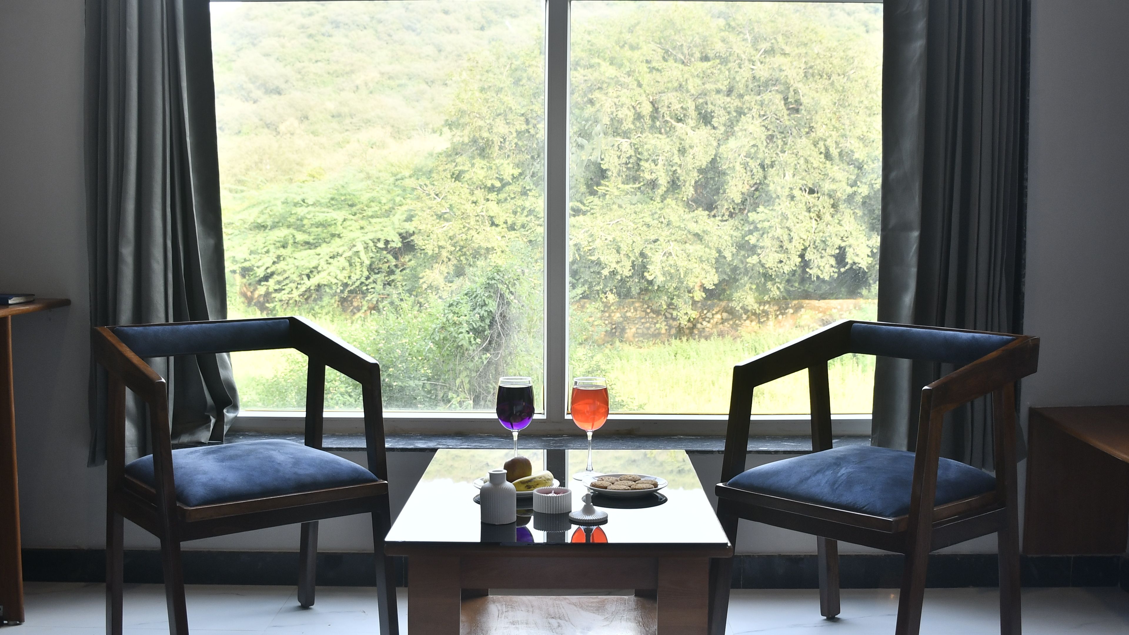 A room with two wooden chairs, a small table, and a window with a view of a forest.