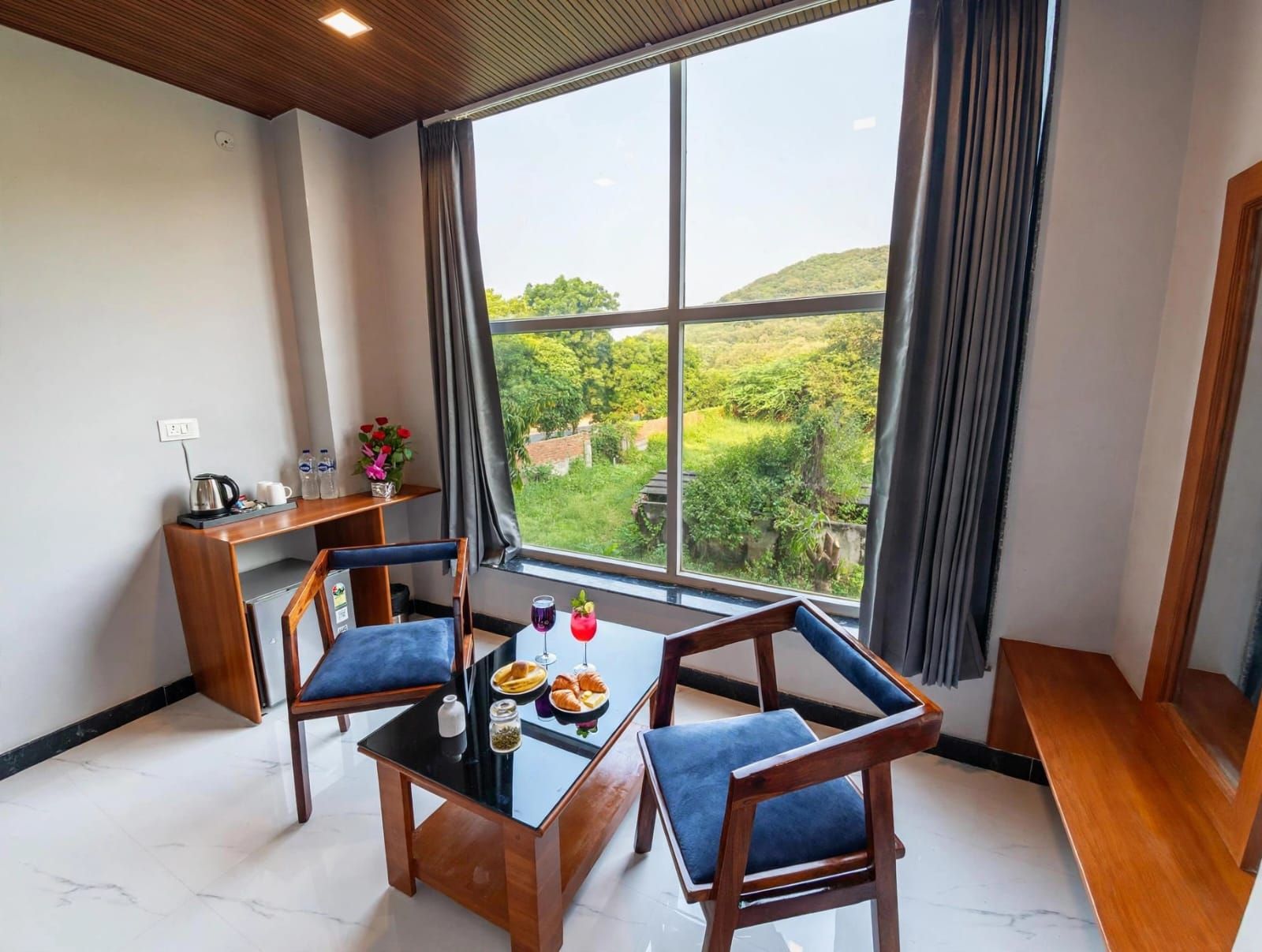 A room with a large window, a table with chairs, a small desk, and a view of greenery outside.