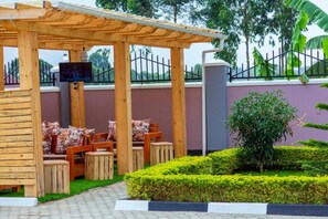 Una pérgola de madera con asientos, una pantalla de televisión y un jardín con setos y un árbol.
