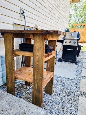 Une table extérieure en bois, avec un banc, un barbecue et un tuyau d’arrosage raccordé à la maison.