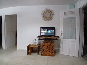 Una habitación con una televisión sobre un soporte de madera, una mesita con un altavoz y una puerta con un panel de vidrio esmerilado.