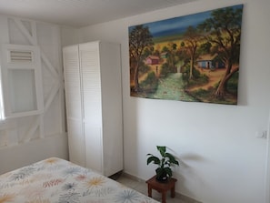 Un dormitorio con una cama, un armario, un cuadro y una mesita con una planta.