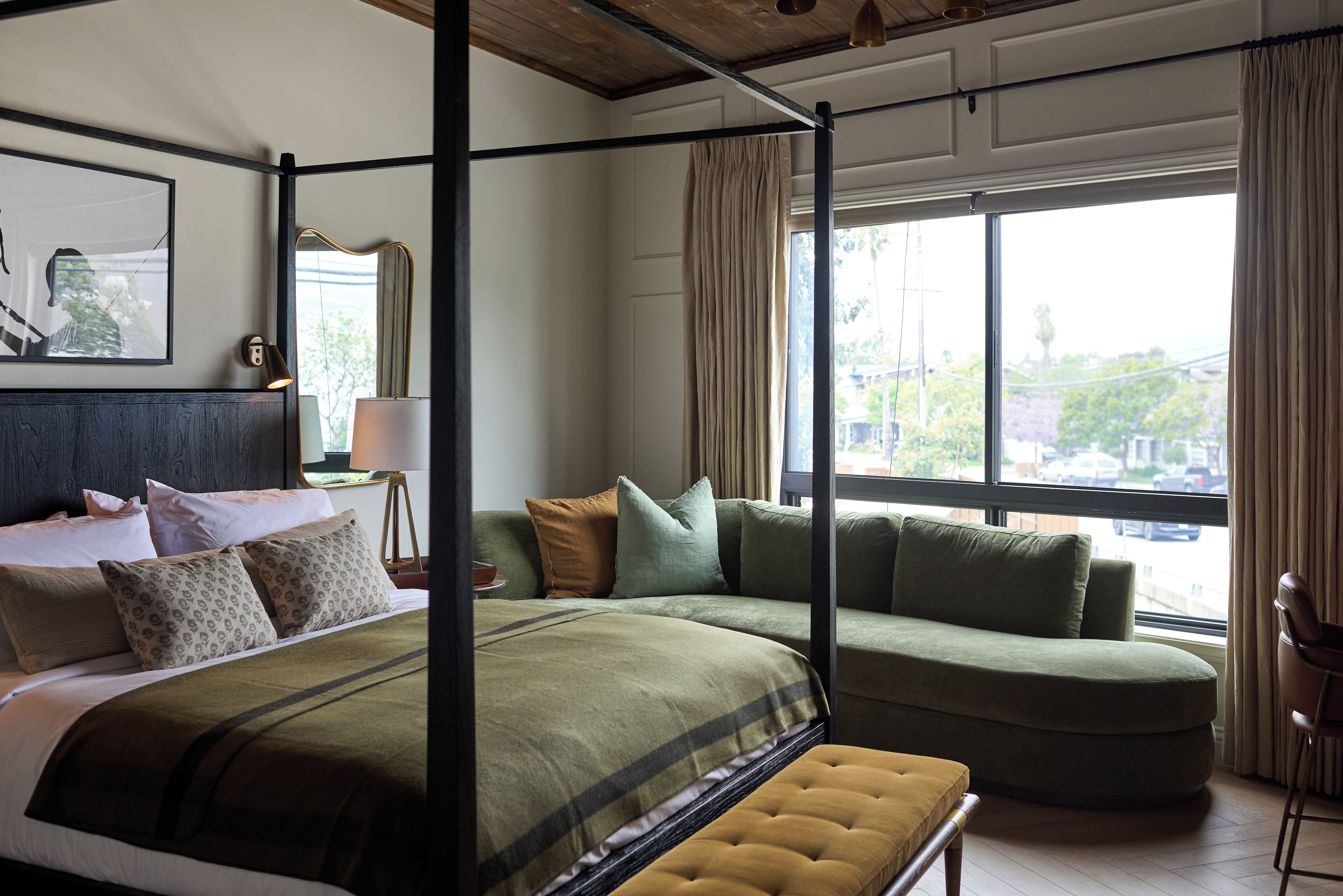 Quarto de hotel com cama de dossel, um sofá, uma espreguiçadeira e uma grande janela com vista para a cidade.
