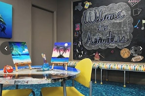 A room with a chalkboard wall displaying 'Welcome to Activities' and various activity icons. There are easels with paintings and a small table with chairs. - Marriott Ocean Pointe Studio w/ King bed, 1 bath, Kitchenette, W/D - Oceanfront (Palm Beach Shores)