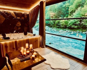 A room with a stone wall, a bathtub, a table with candles, and a view of a river. - AYDER Diamond River (Camlihemsin)
