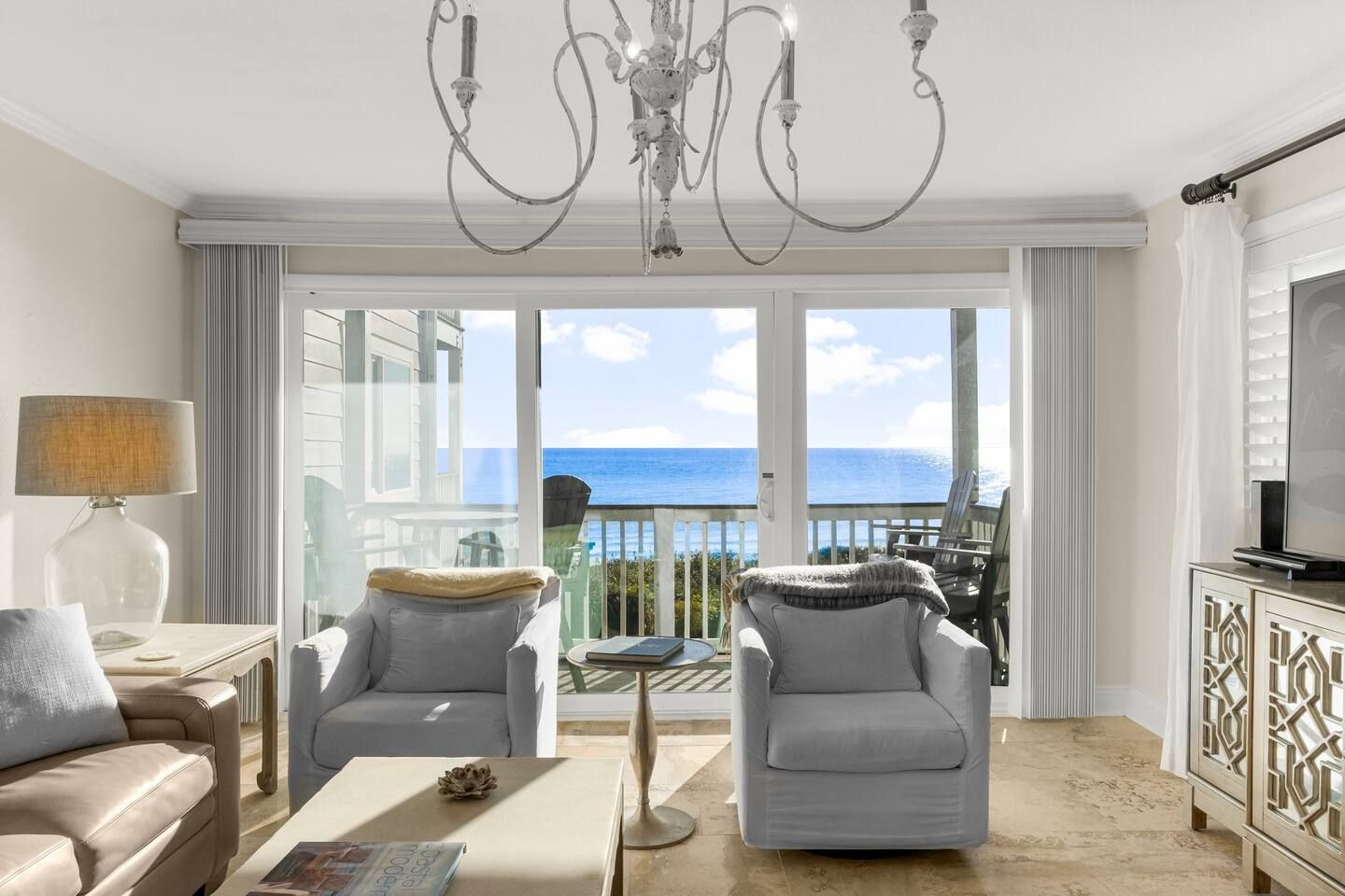 A modern living room with a view of the sea, a chandelier, and a television on a cabinet.